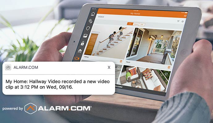 A tablet showing the Alarm.com app with a security alert notification for a hallway video clip