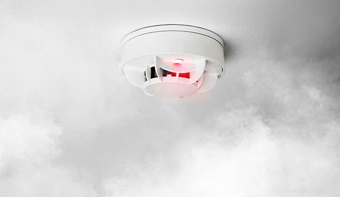 A white smoke detector mounted on a ceiling with a glowing red light as smoke surrounds it