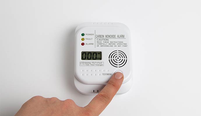 A person pressing the test button on a white carbon monoxide alarm with a digital display showing zero ppm